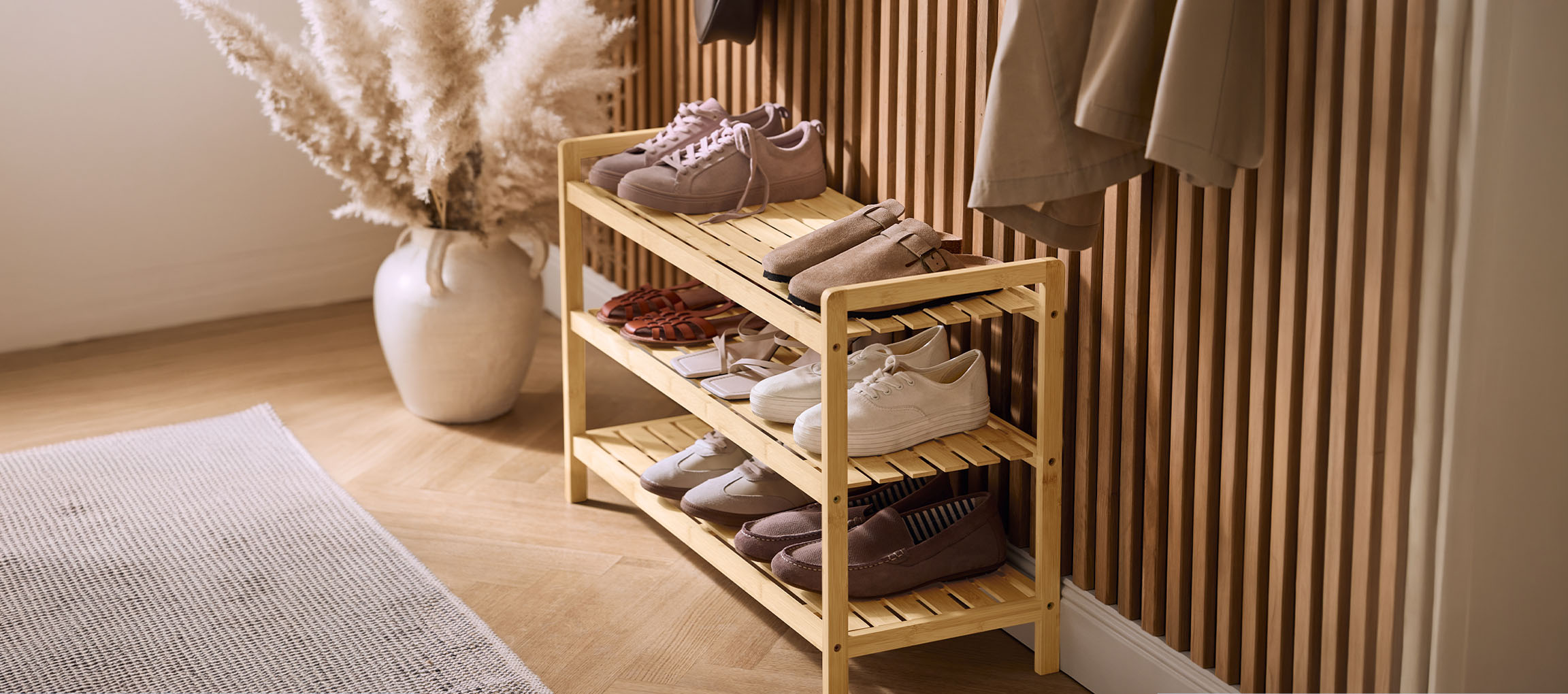 Holzschuhregal mit verschiedenen Schuhen, einer Pampasgrasvase und einem Mantel an der Wand.