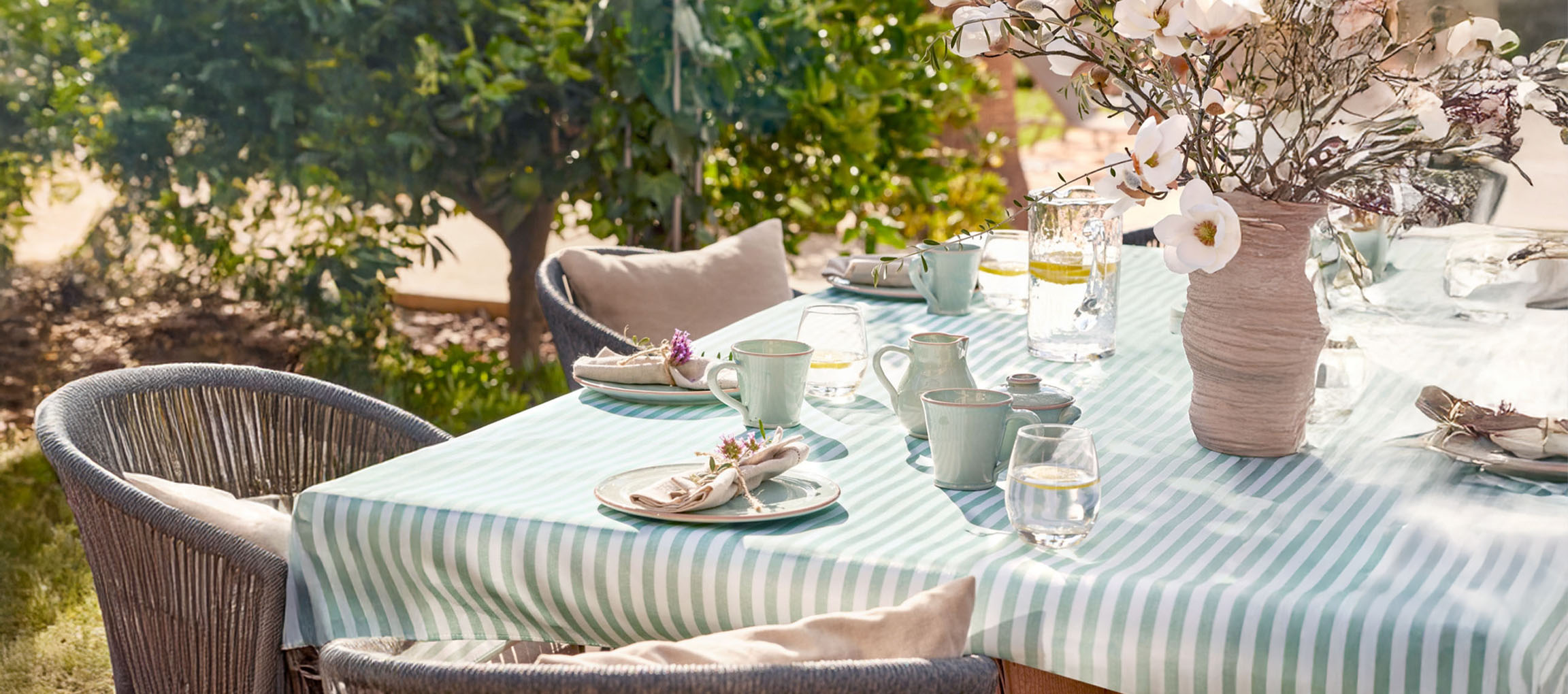 Gedeckter Esstisch im Freien mit gestreifter Tischdecke, mintgrünem Keramikgeschirr und Blumen.