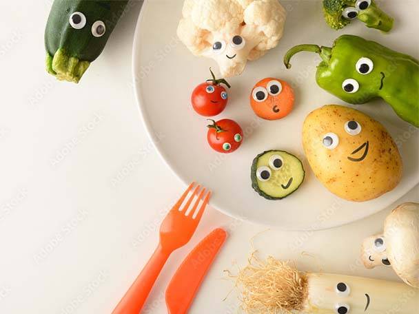 Lustiges Gemüse und Obst mit Kulleraugen auf einem weißen Teller, darunter Zucchini, Blumenkohl und Paprika.