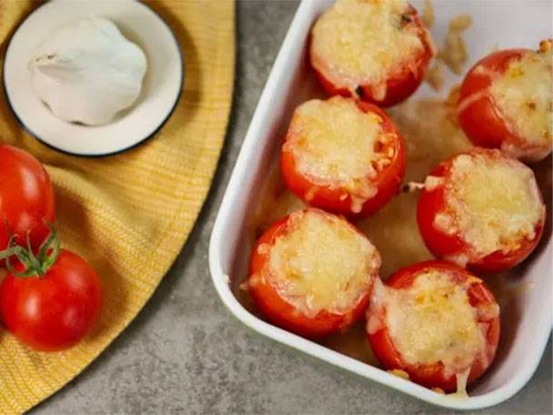Gebackene Tomaten mit Käse in einer Auflaufform, daneben frische Tomaten und Knoblauch.