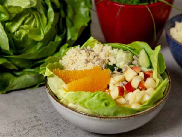 Eine Schüssel mit Salat, Couscous, Orangen, Äpfeln und Gurken auf einem Tisch.