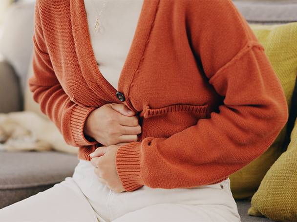 Eine Frau in einem orangefarbenen Strickpullover hält sich den Bauch.