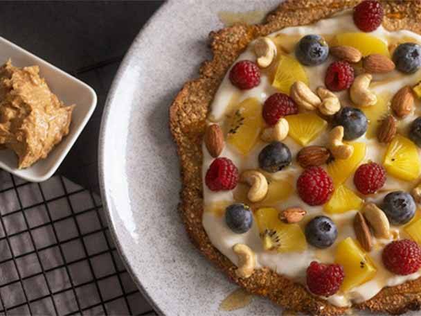 Ein Dessert-Pizza mit Beeren, Nüssen und Früchten auf einem Teller, daneben eine Schüssel mit Erdnussbutter.