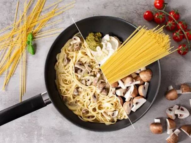 Spaghetti mit Pilzen und Pesto in einer Pfanne, daneben frische Tomaten und rohe Nudeln.