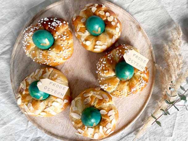 Fünf Osterbrote mit grünen Eiern und „Frohe Ostern“-Anhängern auf einem Holztablett.