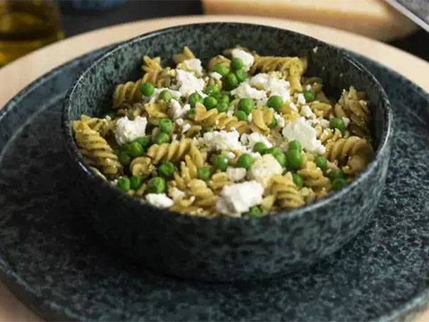 Nudelsalat mit Erbsen und Feta in einer dunklen Schale auf einem Teller.