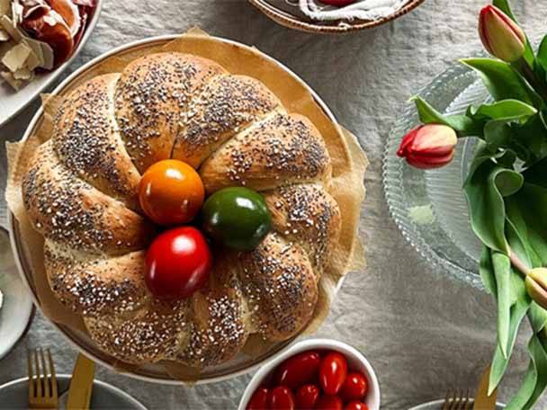 Geflochtenes Brot mit Mohn und bunten Eiern, daneben Tulpen und eine Schale mit Tomaten.