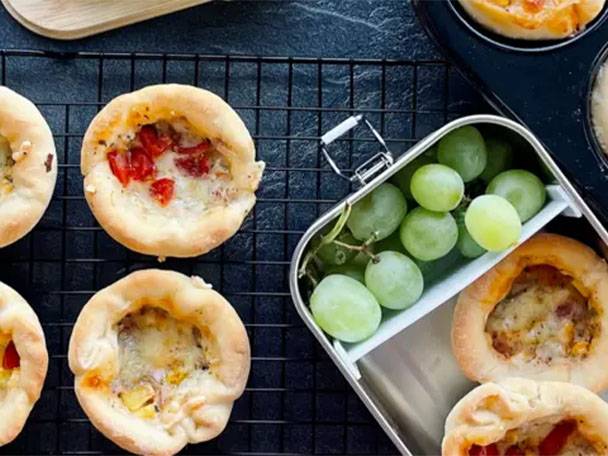 Mini-Pizzen und grüne Weintrauben in einer Brotdose auf einem schwarzen Gitterrost.