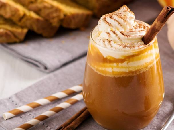 Ein Glas Kürbis-Gewürzgetränk mit Schlagsahne und Zimtstange, daneben Kürbisbrot.