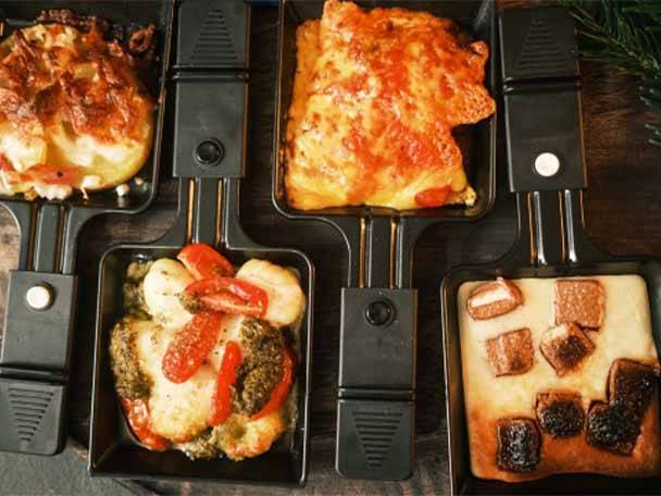 Vier Raclette-Pfännchen mit verschiedenen Gerichten, darunter Kartoffeln, Gemüse und Schokolade.