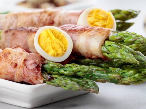 Speck umwickelter Spargel mit hartgekochten Eiern auf einem weißen Teller.