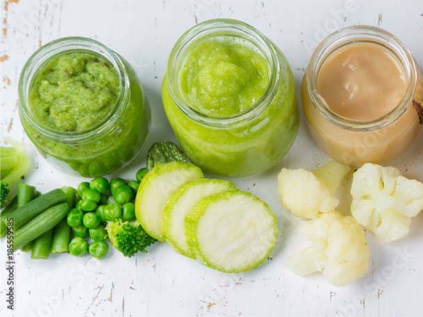 Gläser mit grünem und beigem Babybrei, umgeben von frischem Gemüse wie Erbsen, Zucchini und Blumenkohl.