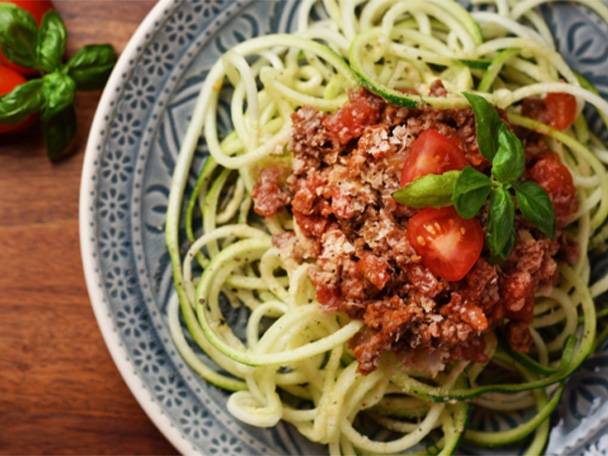Zucchini-Nudeln mit Bolognese-Sauce, Kirschtomaten und Basilikum auf einem Teller.