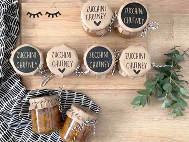 Eine Holzplatte mit Gläsern von Zucchini-Chutney, einige mit schwarzen und einige mit beigen Etiketten.