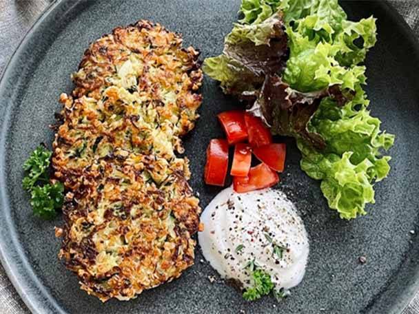 Zwei goldbraune Gemüsepuffer mit Salat, Tomaten und einem Klecks Joghurtsoße auf einem Teller.