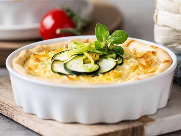 Eine Quiche mit Zucchini und Kräutern in einer weißen Backform auf einem Holzbrett.