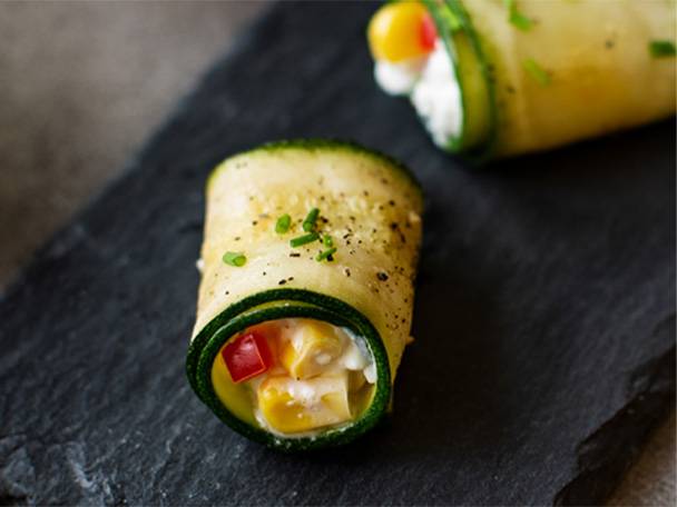 Zwei Zucchiniröllchen mit Frischkäse, Mais und Paprika auf einer dunklen Schieferplatte.