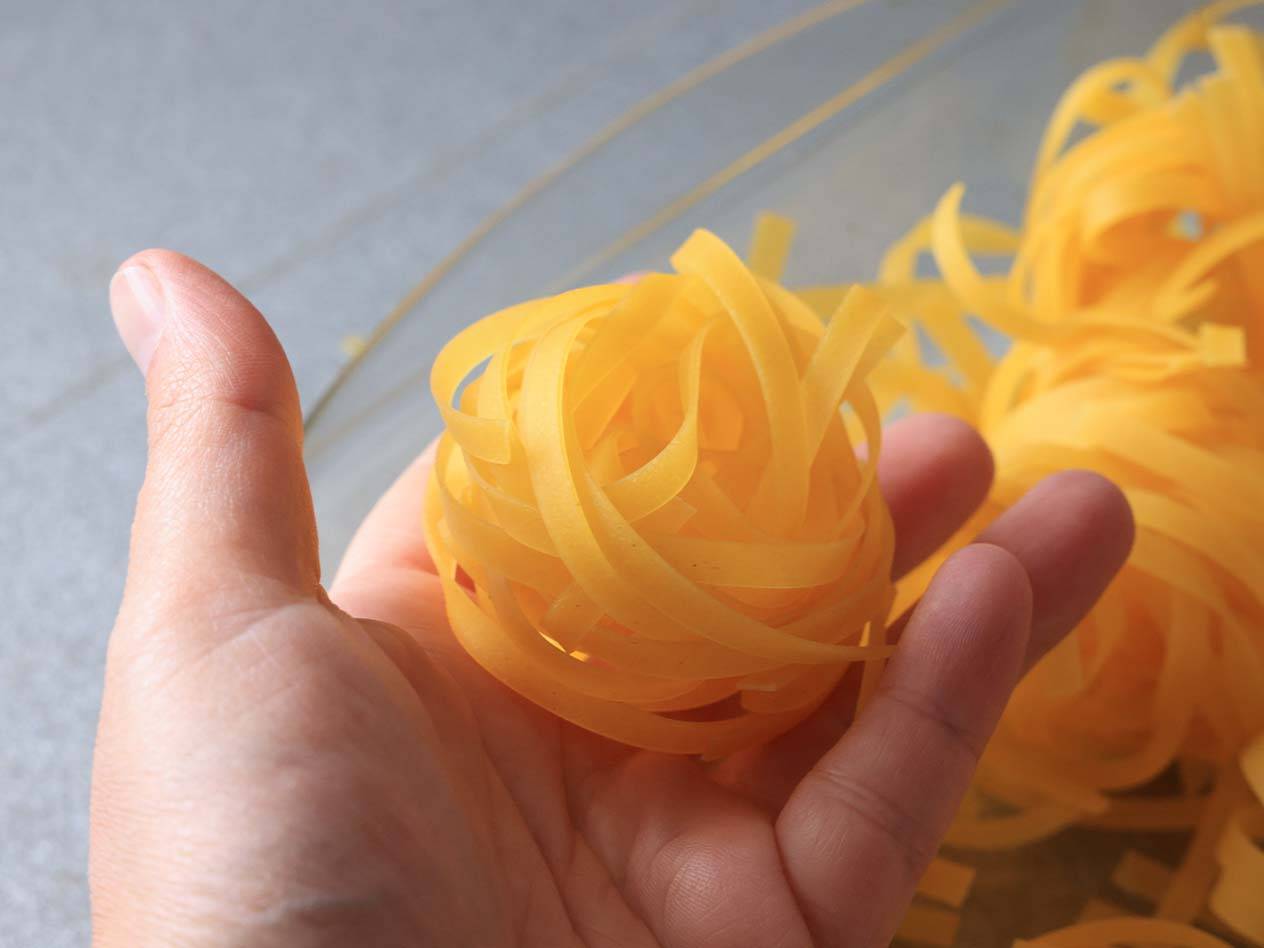 Eine Hand hält ein Nest frischer Tagliatelle über einer Schüssel mit mehr Nudeln.