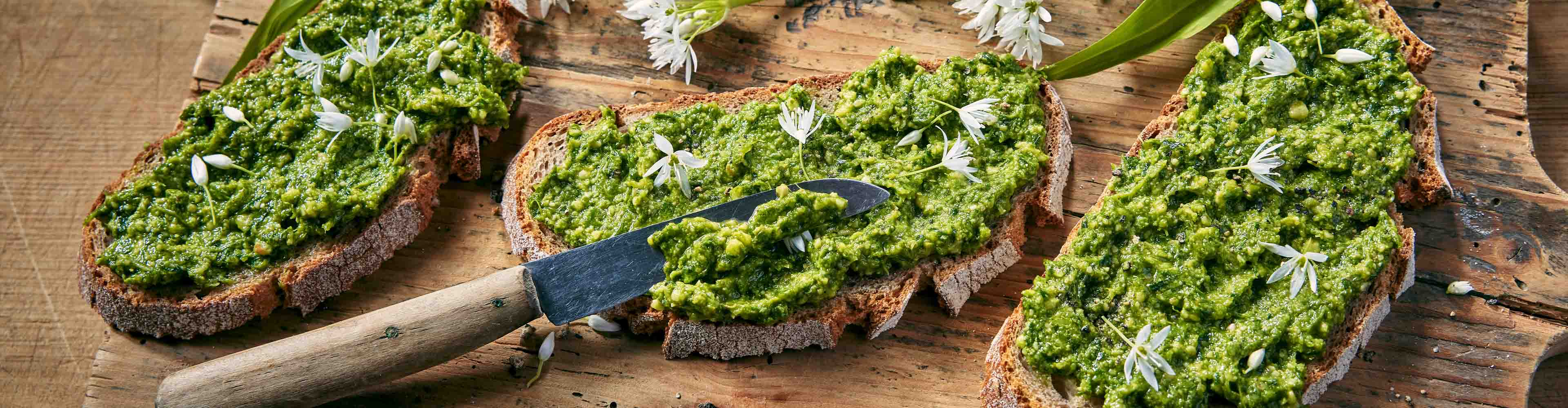Drei Scheiben Brot mit grünem Pesto und weißen Blüten auf einem Holzbrett.