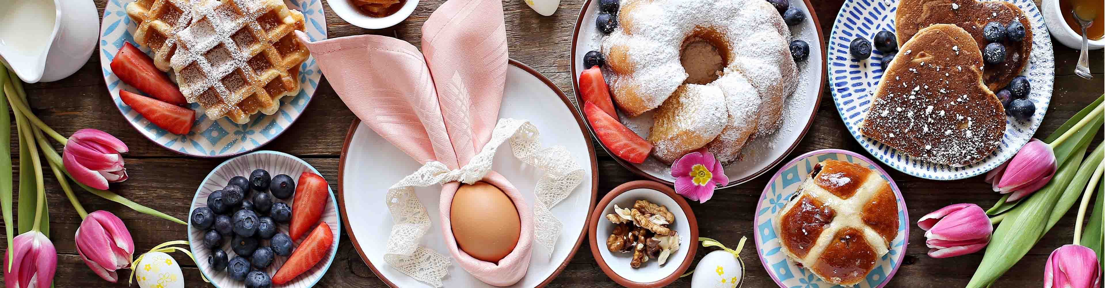 Osterfrühstück mit Waffeln, Kuchen, Pfannkuchen, Hot Cross Buns und frischen Früchten auf einem Holztisch.