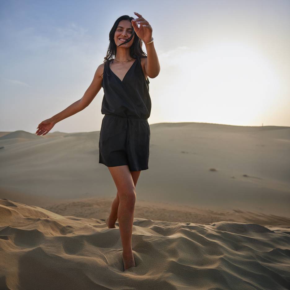 Frau in schwarzem Overall barfuß auf Sanddünen bei Sonnenuntergang.
