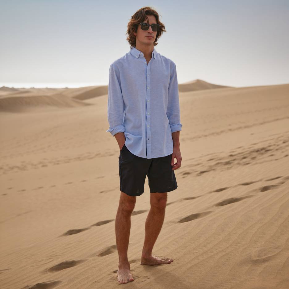 Mann in blauem Hemd und schwarzen Shorts, barfuß auf Sanddünen stehend.