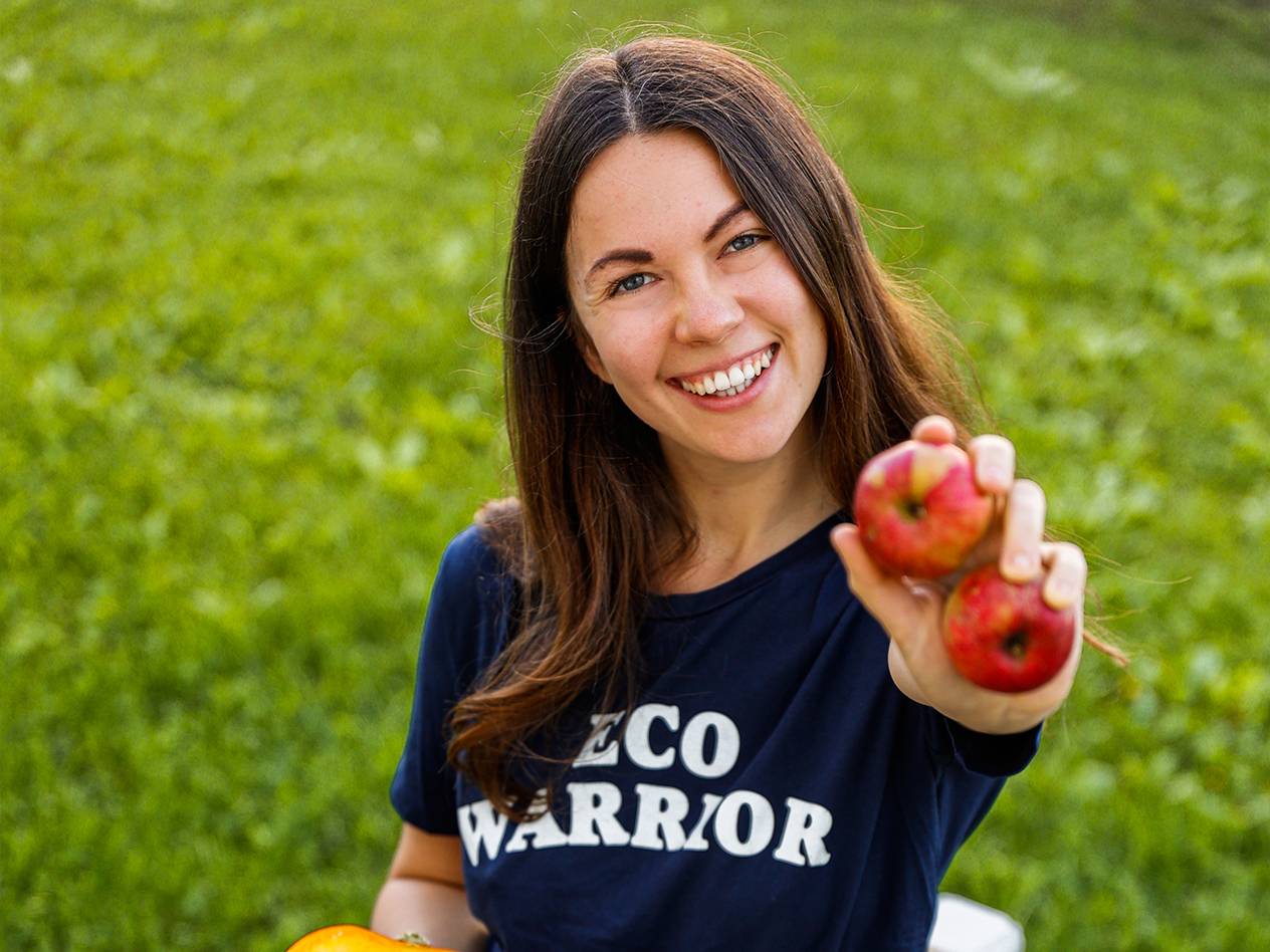 Eine lächelnde Frau in einem 'ECO WARRIOR'-Shirt hält zwei rote Äpfel.