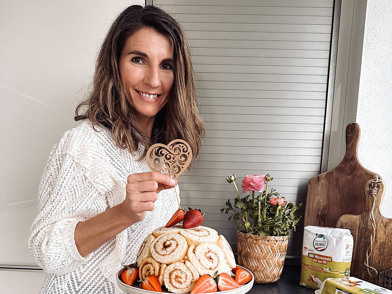 Eine lächelnde Frau hält ein Herz über einem Kuchen mit Erdbeeren und Backzutaten.
