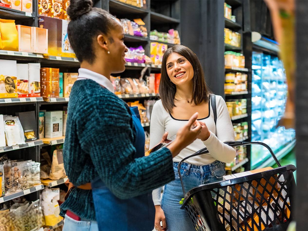 Zwei Frauen unterhalten sich in einem Supermarkt vor Regalen mit Trockenwaren und Getränken.