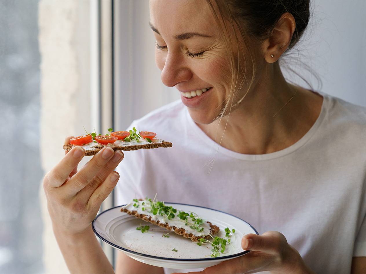 Eine Frau genießt einen gesunden Snack: Knäckebrot mit Frischkäse, Tomaten und Sprossen.