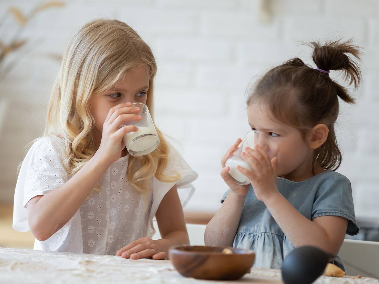 Zwei kleine Mädchen trinken Milch aus Gläsern an einem Tisch.