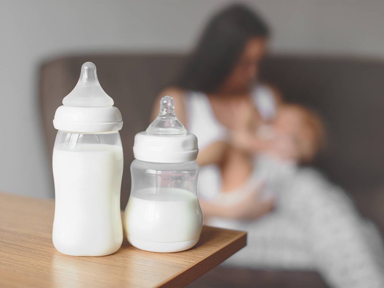Zwei Babyfläschchen mit Milch, im Hintergrund stillt eine Frau ein Baby.