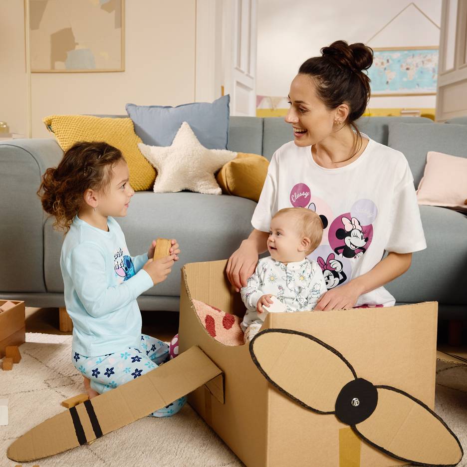 Mutter und Kinder spielen mit einem Karton, die Mutter trägt ein T-Shirt mit Zeichentrickfiguren.