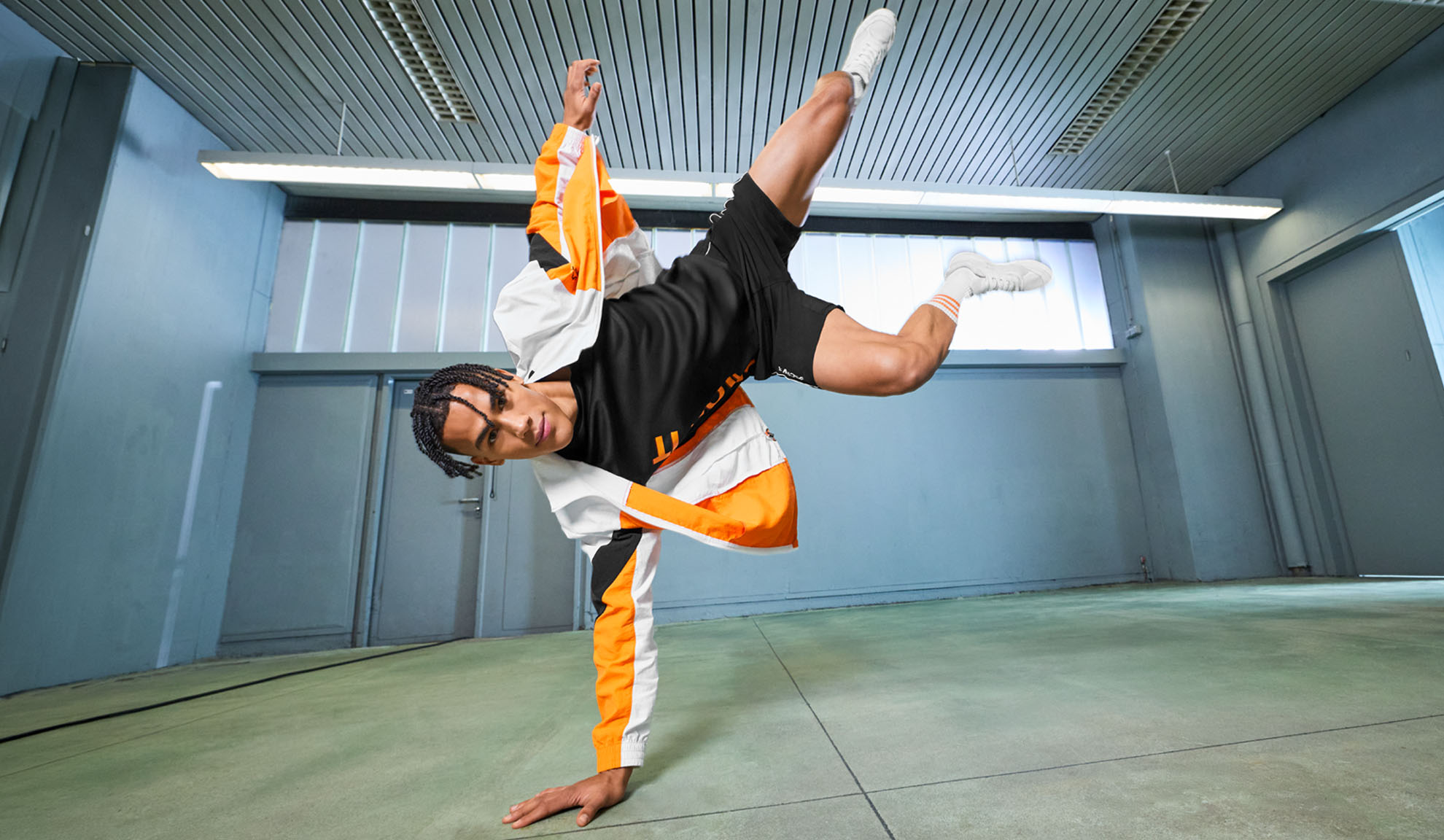 Mann in Sportkleidung, schwarzem T-Shirt, Shorts und orange-weißer Jacke, führt einen Tanzschritt aus.