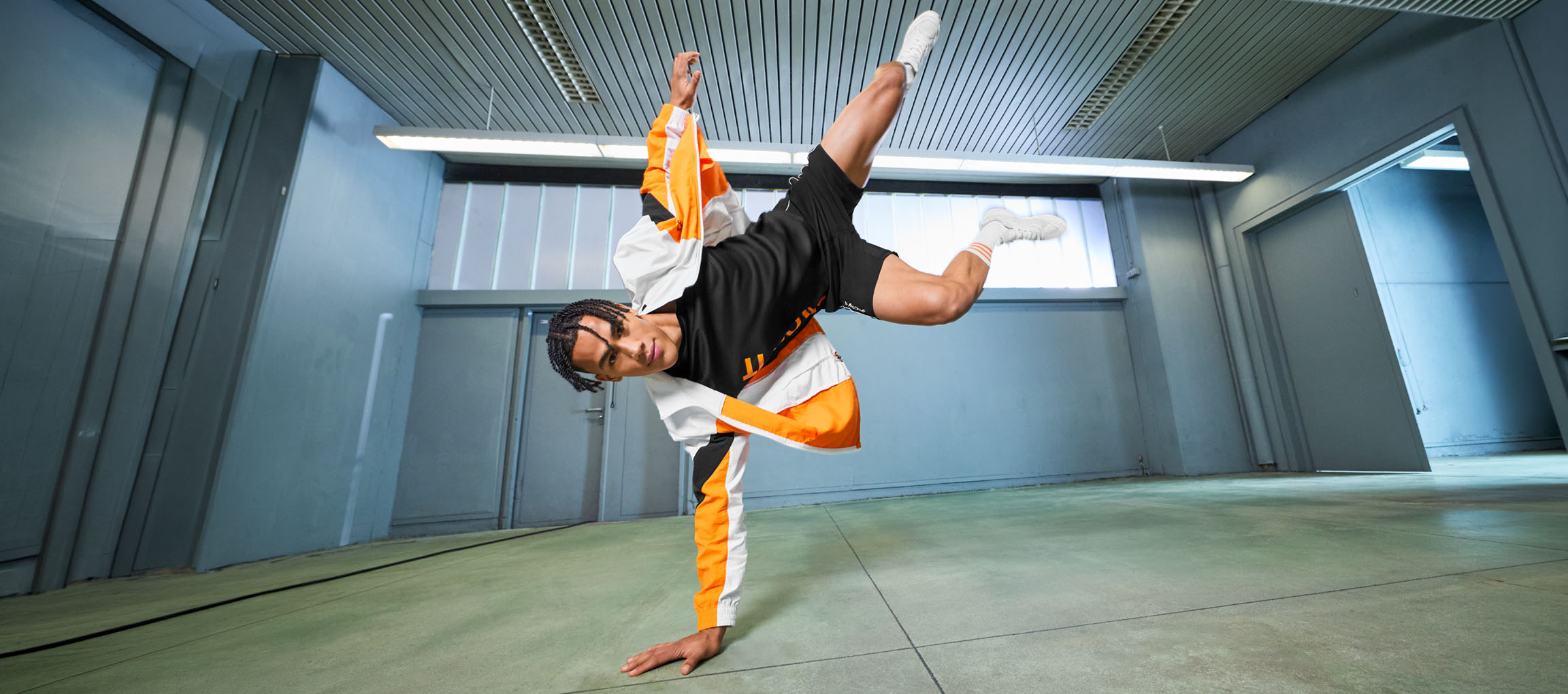 Mann in Sportkleidung, schwarzem T-Shirt und orange-weißer Jacke, tanzt in einem Raum.