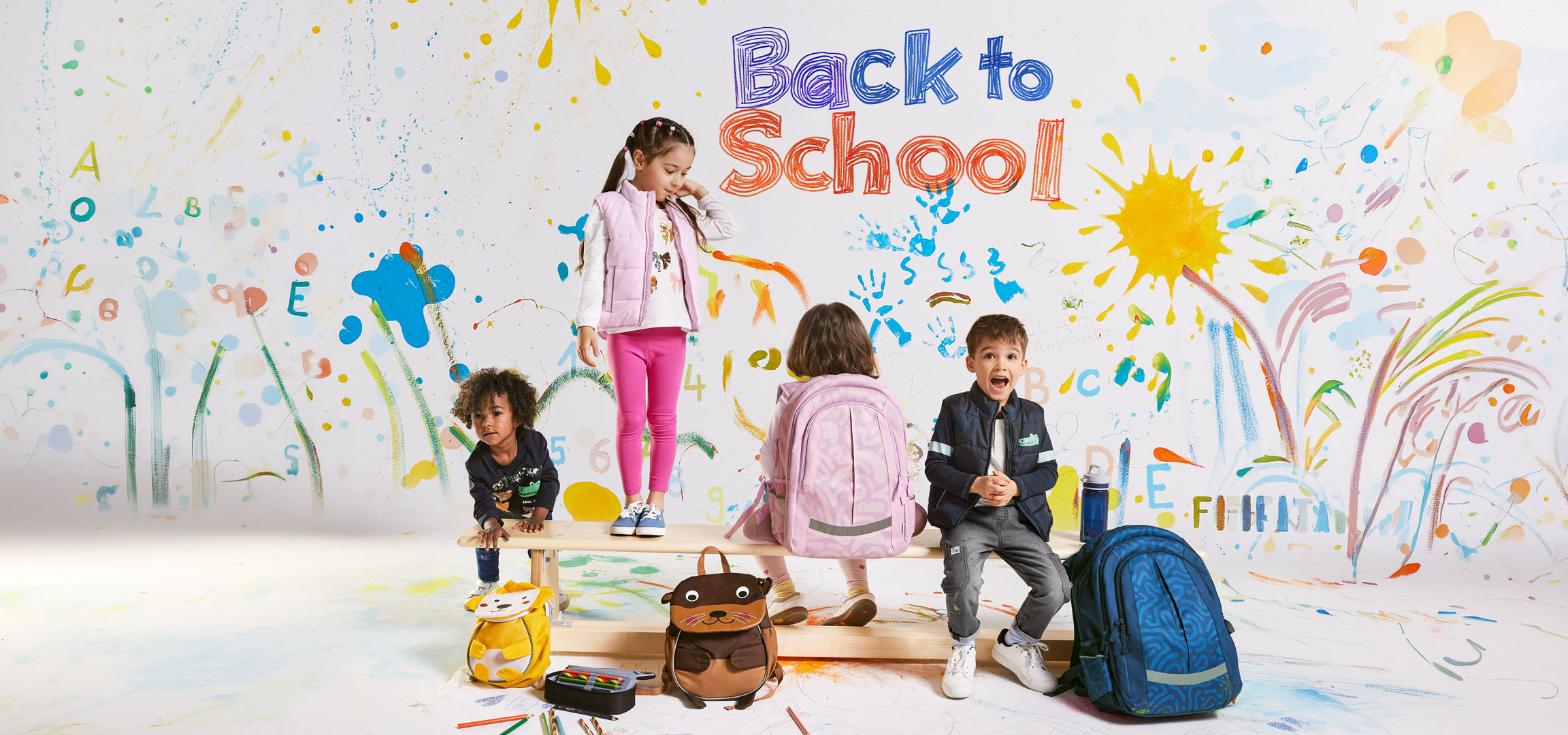 Vier Kinder mit Schulranzen und Rucksäcken vor einer bunten Wand mit der Aufschrift 'Back to School'.