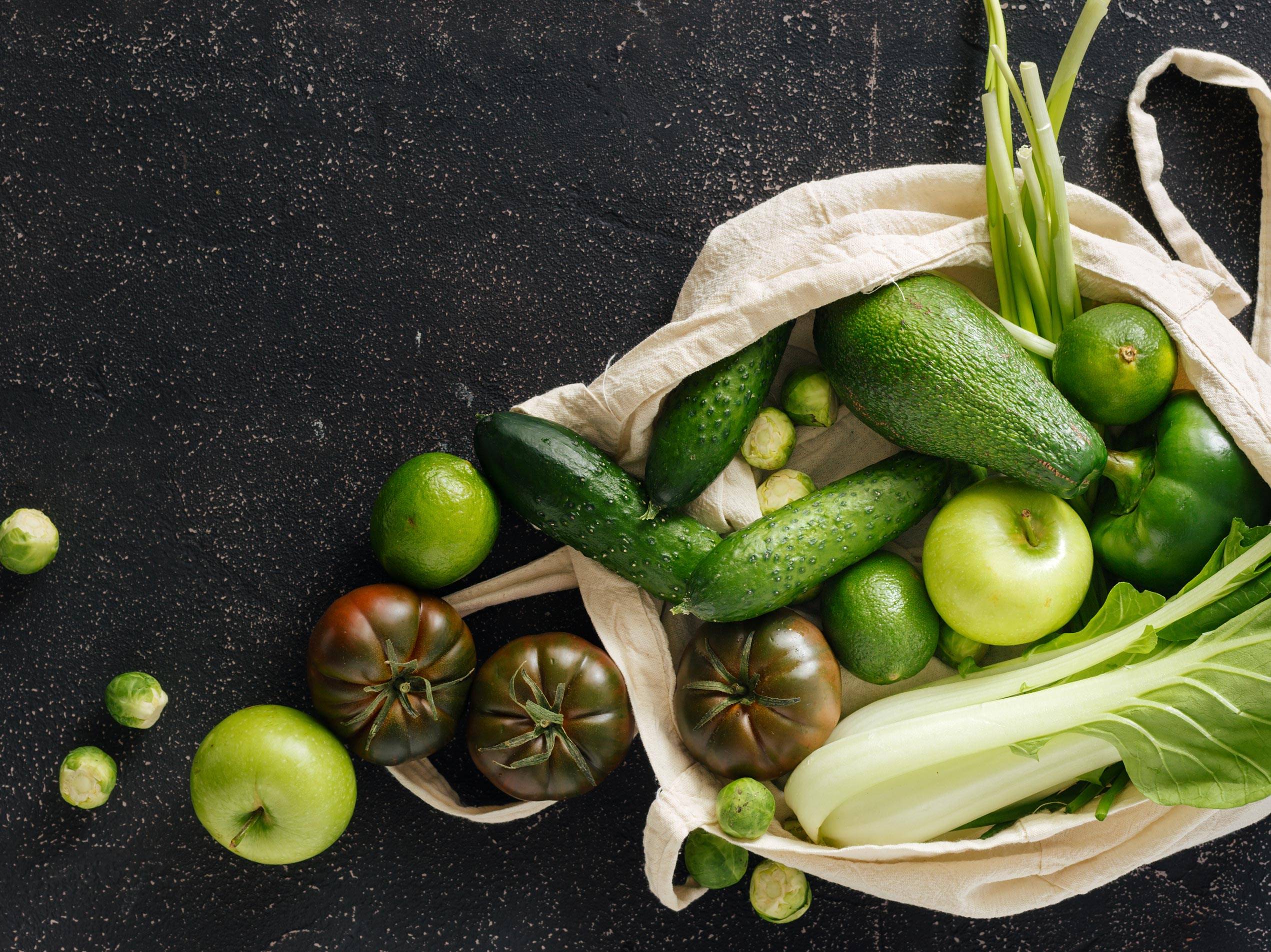 Frisches grünes Gemüse und Obst, darunter Gurken, Avocados, Äpfel und Tomaten, in einer Stofftasche auf dunklem Hintergrund.