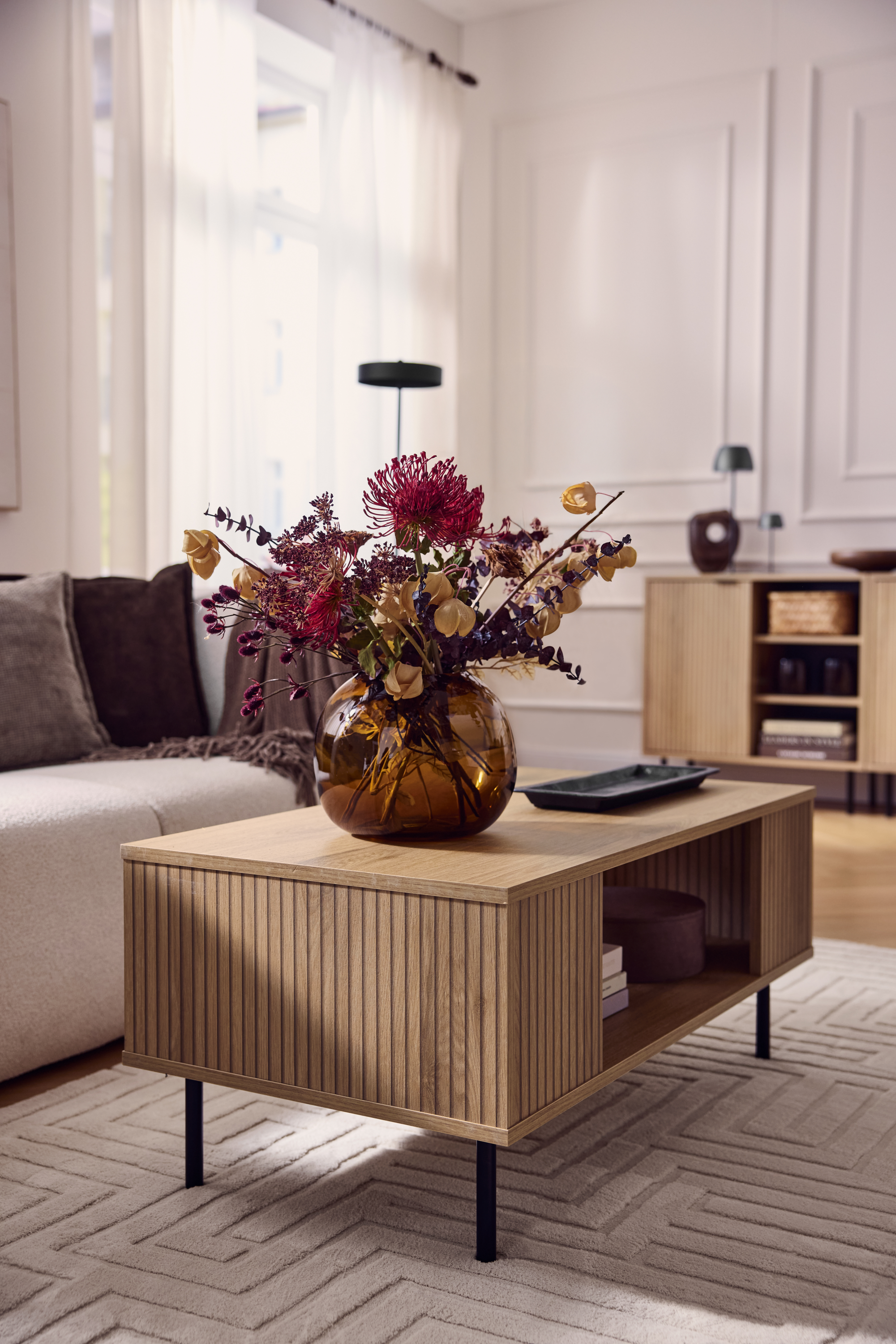 Couchtisch aus Holz mit geriffelter Front, Vase mit Trockenblumen, beiges Sofa.